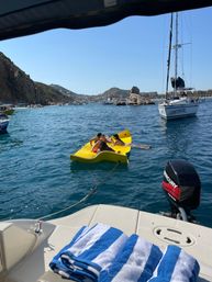 Three people lounging on a bright yellow floating mat tethered to a boat in a sunny coastal bay, surrounded by sailboats, rocky cliffs and a seaside town, with blue-and-white striped towels on the boat deck in the foreground.