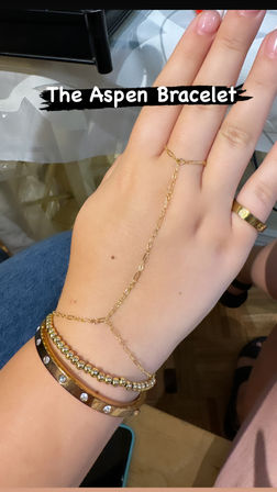 Delicate gold hand-chain bracelet labeled "The Aspen Bracelet" draping from finger to wrist, paired with a gold beaded bracelet and rhinestone-studded bangle on a hand over denim and parquet floor.