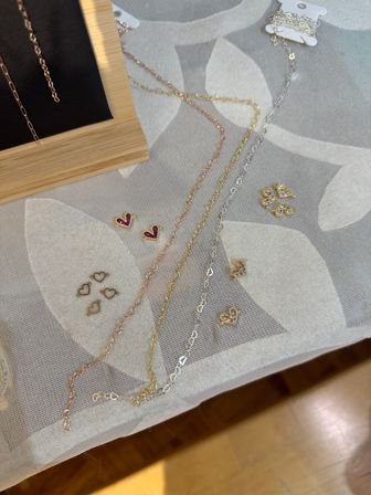 Delicate gold, rose-gold and silver chain necklaces with tiny heart, V and butterfly stud earrings arranged on white leaf-patterned fabric display beside a wooden stand