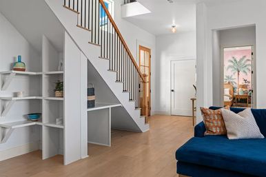 Bright modern living area with an open staircase and black metal balusters, built-in white under-stair shelving, light wood floors, and a blue velvet sofa; doorway shows pink tropical palm wallpaper in a bedroom.