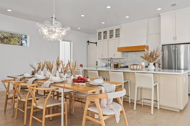 Airy open-concept white kitchen and dining area with a wood farmhouse table, woven chairs, pampas grass centerpieces, bubble pendant light, island with bar stools, and a red fruit centerpiece.