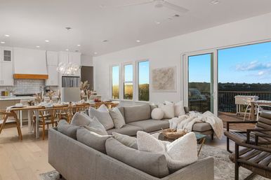 Cozy sunlit modern open-concept living room and kitchen with a gray sectional sofa, wooden dining table, glass pendant lights, light hardwood floors, and sliding doors opening to a balcony with treetop views.