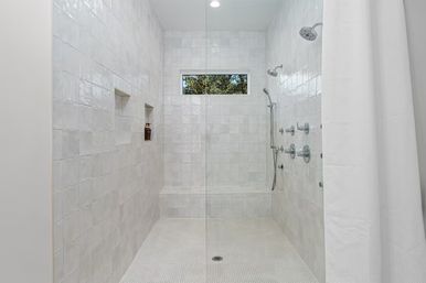 Bright modern white-tiled walk-in shower with glass partition, built-in bench and recessed shampoo niches, multiple chrome showerheads, small high window and textured white floor.