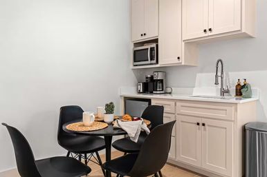Cozy modern kitchenette with white shaker cabinets, stainless microwave and coffee station, gooseneck faucet, and a round black dining table with four black chairs set with mugs, woven placemats and breakfast pastries.