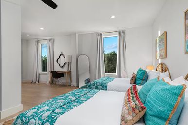 Bright modern guest bedroom with two twin beds, teal patterned duvets and colorful throw pillows, light wood floors, desk and chair, full-length mirror, and tall windows with curtains showing greenery and blue sky.