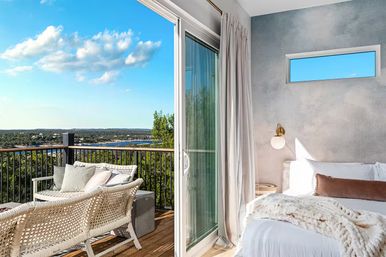 Bright modern bedroom with sliding glass door onto a sunlit balcony featuring wicker seating, overlooking a lake and forest under a clear blue sky — cozy lakeview retreat.
