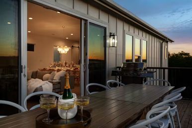Sunset outdoor dining patio on a modern deck with a long wooden table, champagne bottle and coupe glasses on a tray, curved patio chairs, sliding glass doors to a warmly lit living and dining room, and a gas grill.