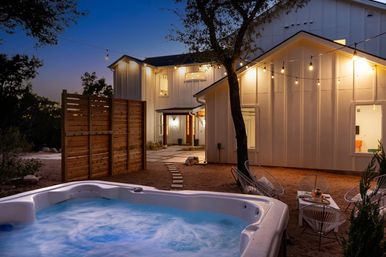 Cozy modern farmhouse backyard at dusk with bubbling outdoor hot tub in foreground, string lights overhead, wood privacy screen, patio seating and illuminated white-siding house.