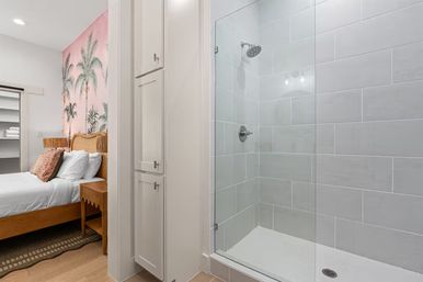 Bright coastal-style bedroom with pink palm wallpaper, wooden bed and nightstand, and a glass-enclosed white-tiled en-suite shower.