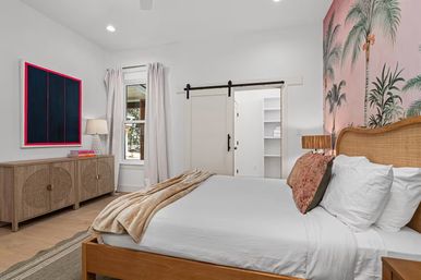 Coastal-style master bedroom with rattan headboard, king bed in white linens and beige throw, pink palm wallpaper accent wall, wood dresser, window with curtains and sliding barn-door closet.