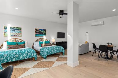 Bright coastal-inspired open-plan studio with two twin beds in teal floral bedding and rattan headboards, jute area rug, beachy wall art, wall-mounted TV, gray sofa, black dining set, ceiling fan and split air conditioner