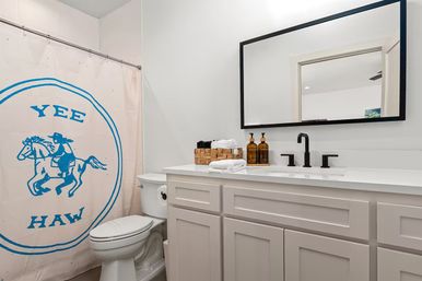 Bright modern bathroom with white shaker vanity, black matte faucet and framed mirror, toilet, and playful pink shower curtain featuring a blue cowboy "YEE HAW" graphic.