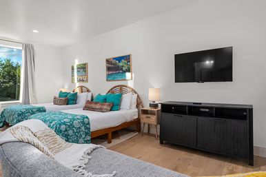 Coastal-style vacation rental bedroom with two double beds, teal patterned throws, rattan headboards, colorful pillows, bedside lamp, wall-mounted TV and large window with tree views.