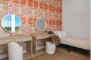Bright modern boho bedroom interior with bold pink-orange patterned wallpaper, two light wood vanities topped with round mirrors reflecting a sunny window view, a striped twin bed, cream boucle pouf with a faux-fur throw, and a jute rug on light wood floors.