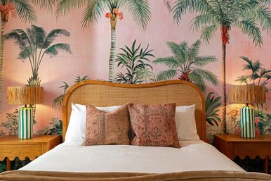 Tropical-chic bedroom with pink palm wallpaper, curved rattan headboard, white bedding, textured terracotta throw pillows, wooden nightstands and green-and-white striped lamps
