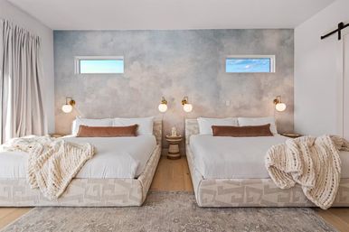 Modern coastal-style bedroom with two matching low-profile platform beds dressed in white linens and brown lumbar pillows, chunky cream knit throws, textured blue-gray accent wall with two narrow sky-view windows, brass globe wall sconces, round woven nightstand, light wood floor and neutral area rug.