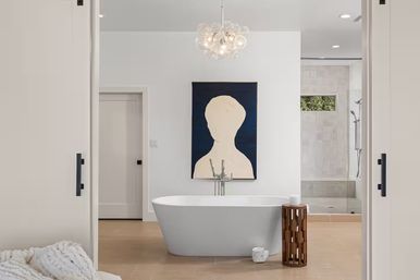 Spa-like modern master bathroom with a white freestanding bathtub centered beneath an abstract portrait, bubble pendant chandelier, wooden accent side table, beige floor tiles and a glass walk-in shower in the background.