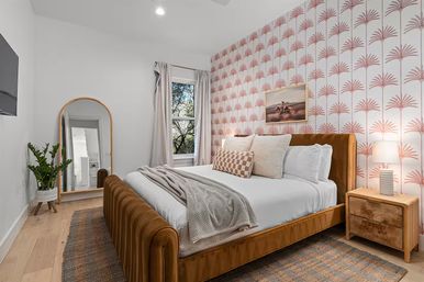 Cozy modern bedroom interior with rust velvet platform bed, pink palm-pattern wallpaper, wooden nightstand and lamp, arched full-length mirror, potted plant, and window view of trees.
