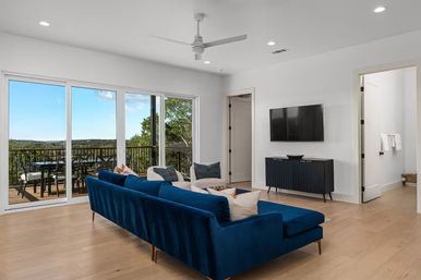 Bright modern living room with deep-blue velvet sectional, light hardwood floors, wall-mounted TV and ceiling fan; large sliding glass doors open to a balcony with outdoor dining and scenic treetop views.