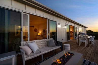 Sunset-lit wooden deck with woven sofa and cushions, rectangular fire pit, outdoor dining table and chairs, and sliding glass doors opening to a warm interior.