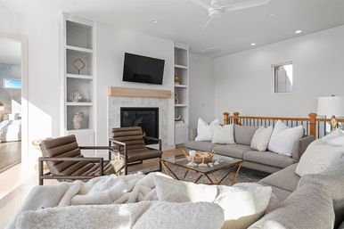 Bright modern living room with cozy gray sectional, two leather lounge chairs, glass-and-brass coffee table, fireplace with wooden mantel, wall-mounted TV, built-in shelves, white walls and ceiling fan.