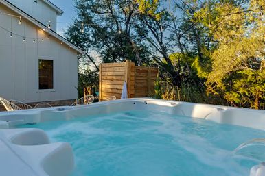 Bubbling outdoor hot tub on a backyard patio with string lights, wooden privacy screen and relaxed wooded golden-hour setting.