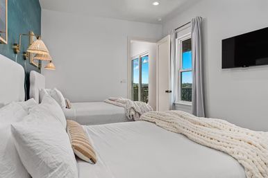Bright modern twin bedroom with crisp white linens, chunky knit throw, rattan wall sconces and a wall-mounted TV, with an open door to a balcony showing blue sky and treetop views, ideal for a coastal-style vacation rental.
