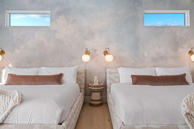 Serene modern coastal-inspired bedroom with two matching beds, neutral textured plaster wall, brass globe sconces, rust velvet lumbar pillows, chunky knit throws and high horizontal windows showing blue sky.