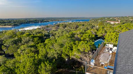 Rooftop terrace with a long outdoor dining table and lounge seating overlooking dense green trees and a blue lake marina with docks under a clear sky.