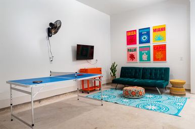 Vibrant indoor rec room with blue ping-pong table, teal velvet sofa, patterned turquoise rug, round pouf, wall-mounted TV, cactus and colorful travel-style posters.