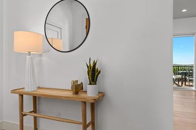 Modern bright entryway with a wooden console table topped by a tall white lamp, brass bird decor and potted snake plant under a round black mirror; sliding door reveals balcony with outdoor seating and distant landscape view.