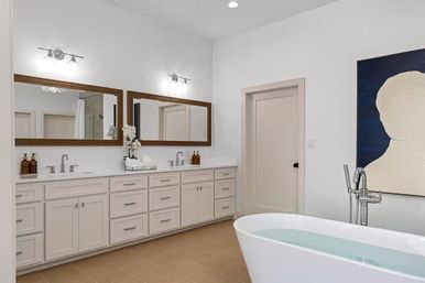 Bright spa-like modern bathroom with long white double-sink vanity, wood-framed mirrors and wall sconces, chrome faucets and soap dispensers, neutral tile floor, minimalist wall art, and a freestanding soaking tub partially filled with water.