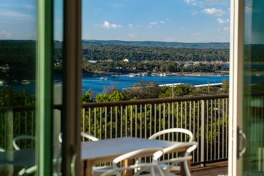 Sunny lake-view balcony with table and white chairs seen through sliding glass doors, overlooking blue water with boats, forested shoreline and rolling hills.