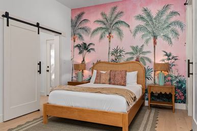 Cozy tropical bedroom with a wooden platform bed and rattan headboard against a pink palm tree mural accent wall, patterned pillows, matching wooden nightstands with striped lamps, and a white sliding barn door.