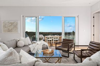 Bright modern coastal living room with light-gray sectional, plush pillows, leather lounge chairs and glass coffee table, sliding glass doors opening to a sunny balcony dining set and sweeping ocean horizon view.