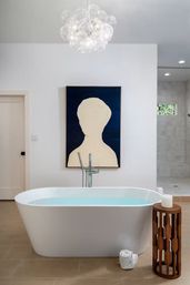 Spa-like modern bathroom in a contemporary home featuring a white freestanding tub with pale blue water, abstract silhouette artwork on a white wall, bubble chandelier overhead, wooden side table with a candle and rolled towel, neutral tile floor and glass walk-in shower.