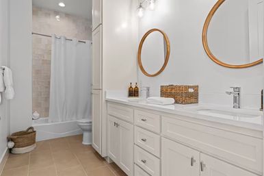 Bright modern residential bathroom with white double-sink vanity, round wood-framed mirrors, woven storage basket, beige floor tiles and shower-tub with white curtain.