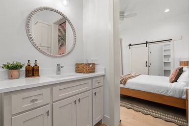 Bright modern en suite bathroom with white vanity, round beaded mirror, amber soap dispensers and wicker basket, opening to airy bedroom with wooden platform bed and black sliding barn door.