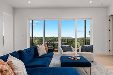 Bright modern living room with deep blue velvet sectional, two white lounge chairs and ottoman with a chess set, sliding glass doors opening to a balcony overlooking treetops and rolling hills under a blue sky.