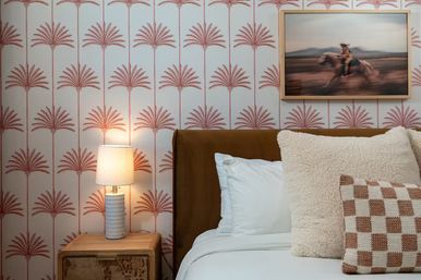 Cozy boho bedroom corner with pink palm-print wallpaper, wooden nightstand and lit ceramic lamp, tan leather headboard, crisp white bedding and textured throw pillows, framed blurred horseback art.