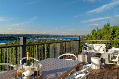 Sunlit waterfront deck with a wooden dining table set with champagne and coupe glasses, white lounge seating and a fire table, railing overlooking a blue bay and tree-lined shoreline under a clear sky.