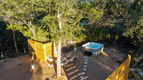 Cozy wooded backyard hot tub oasis with privacy wooden screens, modern lounge chairs, potted plants, string lights, and a stepping-stone path