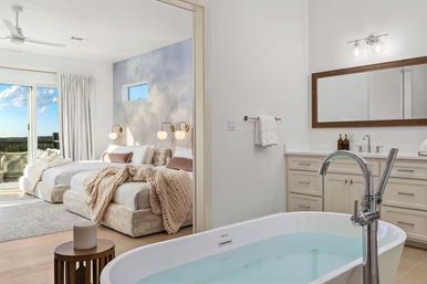 Bright luxury master suite with open ensuite: freestanding soaking tub and chrome floor faucet, double vanity, cozy beds with chunky knit throws, globe sconces and balcony with scenic view.
