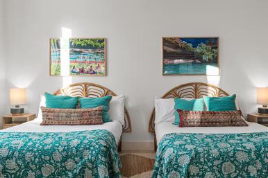 Sunlit twin bedroom with rattan headboards, teal patterned quilts and pillows, decorative lumbar cushions, wooden nightstands with lamps, and colorful coastal paintings on white walls.