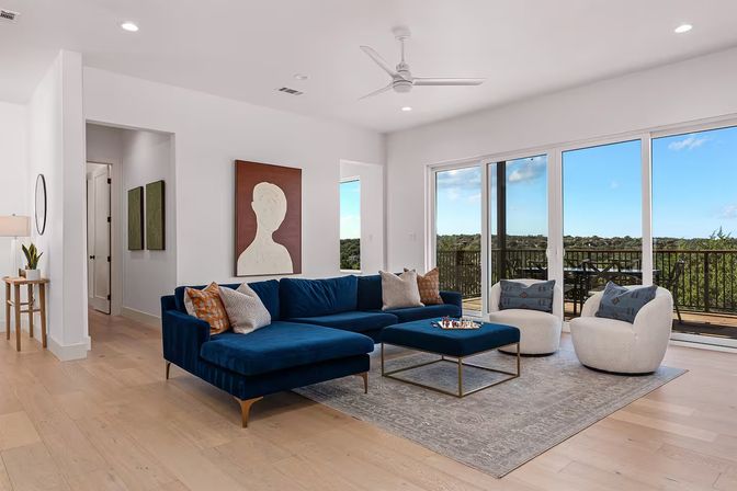 Sunlit modern living room with a navy velvet sectional, cozy white swivel chairs, hardwood floors and sliding glass doors opening to a balcony with treetop views.