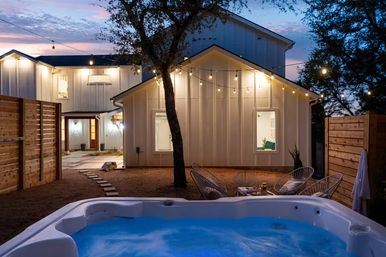 Cozy modern farmhouse backyard at dusk with glowing hot tub in the foreground, string lights draped between trees, white siding house, patio seating and wooden privacy fence.