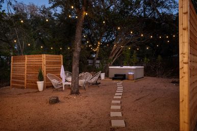 Cozy backyard patio at dusk with warm string lights, stepping stones leading to a hot tub, white woven lounge chairs around a small table, wooden privacy screens and potted plants.