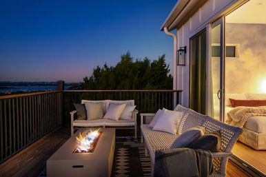 Cozy evening outdoor deck with wicker sofas and cushions around a rectangular fire pit table, sliding glass doors opening to a lit bedroom and a distant water view at dusk.