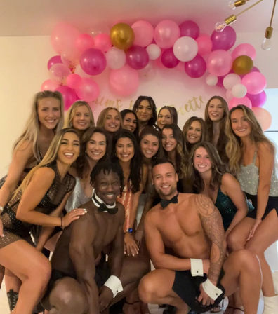 Smiling group of women gathered with two shirtless male entertainers in bow ties, posing under a pink and gold balloon arch at an indoor bachelorette party celebration.