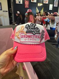 Bright pink trucker hat with patches reading "a lil bougie" and "BRUNETTES do it better" held up at an indoor girls' hat-decor party with kids in caps and wall art in the background.